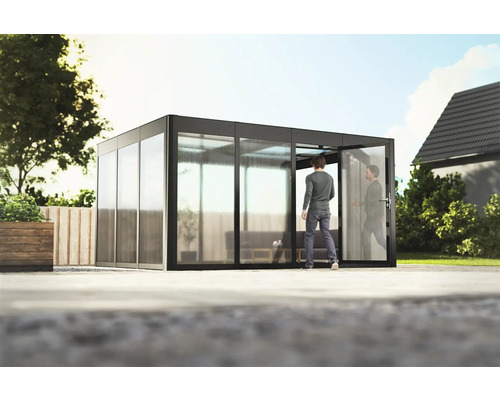 Gewächshaus SunGarden Select 4.3 Polycarbonat 10 mm 443 x 338 cm anthrazit Außenansicht eines modernen Gartenhauses mit Glasschiebetüren und einer Person, die hineingeht.