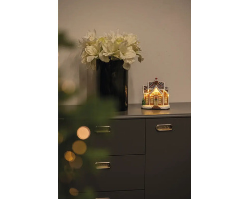 Dekorative Szene mit Vase mit Kunstblumen und beleuchtetem Lebkuchenhaus auf einer Kommode.