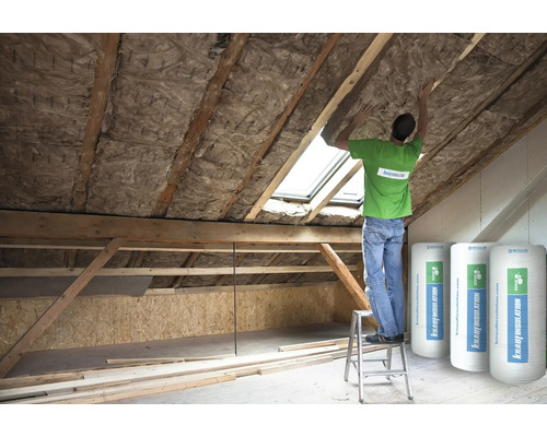 Ein Mann isoliert ein Dachgeschoss mit Dämmwolle. Im Raum stehen Rollen mit Knauf Insulation Logo.