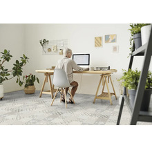 Eine Frau sitzt an einem Schreibtisch mit einem Computer in einem hellen Raum mit Pflanzen und gemustertem Bodenbelag.