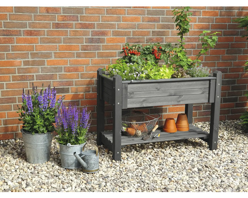 Hochbeet aus Holz mit Pflanzen und Gartenutensilien vor einer Ziegelwand