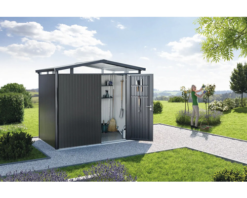 Gartenschuppen mit geöffneter Tür in einer Gartenlandschaft mit Rasen und einer Frau, die Rosen beschneidet.