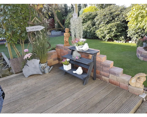 Gartenszene mit Holzterrasse, Blumen und Dekorationen wie Vogeltränken, Figuren und Pflanzen