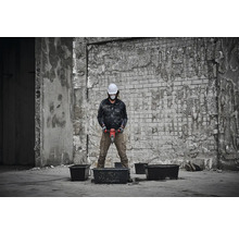 Eine Person mit Schutzhelm und Schutzmaske mischt Material mit einem Rührwerk in einer Bauumgebung.