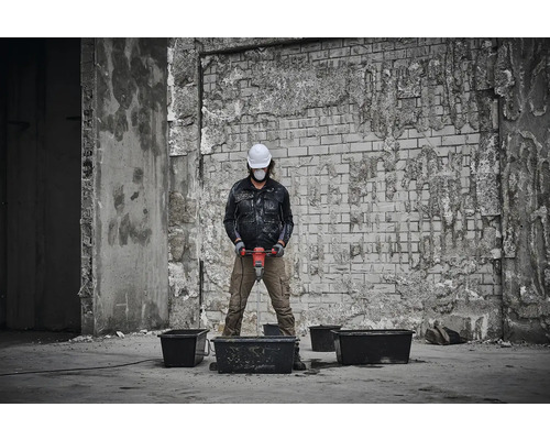 Eine Person mit Schutzhelm und Schutzmaske mischt Material mit einem Rührwerk in einer Bauumgebung.