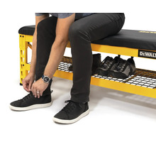 Eine Person sitzt auf einer gelben Werkbank mit Schuhablage und schnürt ihre Schuhe. Auf der Werkbank befinden sich weitere Schuhe. DeWalt Logo