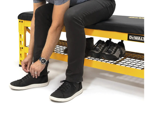 Eine Person sitzt auf einer gelben Werkbank mit Schuhablage und schnürt ihre Schuhe. Auf der Werkbank befinden sich weitere Schuhe. DeWalt Logo