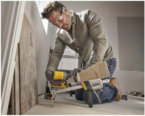 Ein Mann kniet und arbeitet mit einem Akku-Schlagschrauber und einer Schraubzwinge an einem Holzbrett