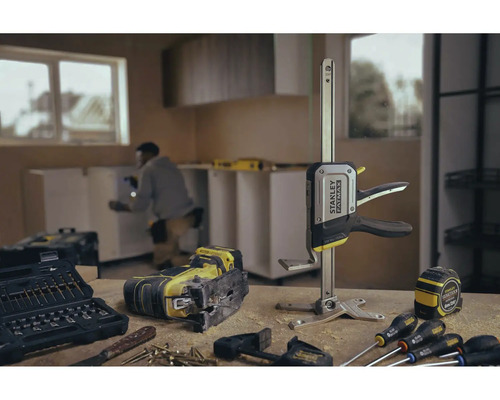 Werkbank mit Stichsäge, Werkzeugkasten, Schraubzwinge, Maßband und Schraubenziehern vor einem Raum mit Küchenzeile.