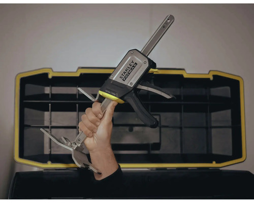 Eine Hand hält eine Stanley Fatmax Einhandzwinge vor einer Werkzeugkiste.