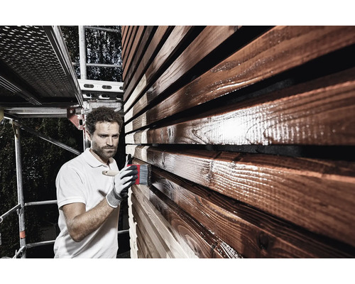 Mann streicht Holzfassade mit Pinsel und Holzschutzlasur von einem Gerüst aus