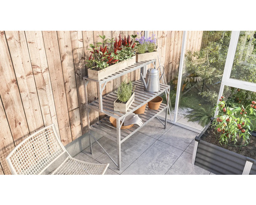 Gartenszene mit Pflanztisch, Pflanzen und Gartengeräten auf einer Terrasse neben einem Gewächshaus