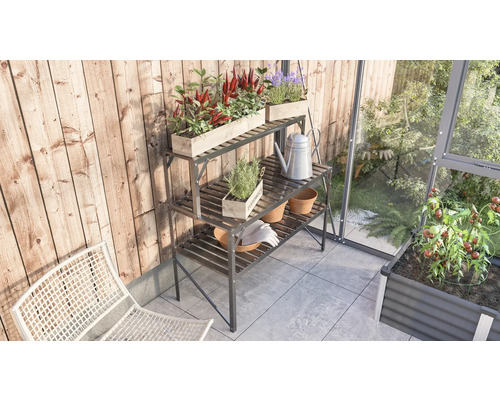 Drei Etagen Pflanztisch aus Metall mit Pflanzen, Töpfen, Gießkanne und Gartenhandschuhen auf einem Balkon neben einem Gewächshaus.