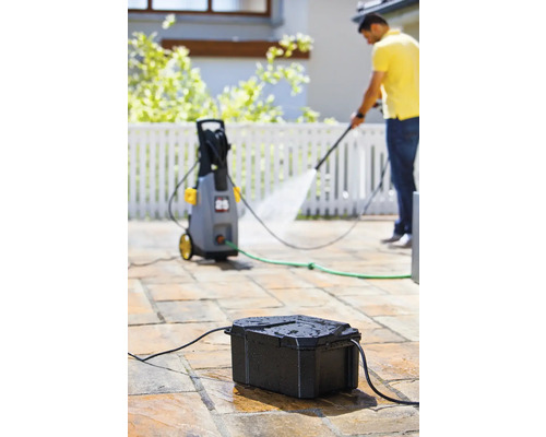 Szene mit Hochdruckreiniger, Wasseranschlusskasten und Person bei Reinigungsarbeiten im Freien.