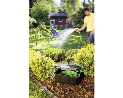 Gartenszene mit versenkter Schlauchbox, Gartenschlauch und einer Person, die mit einer Gartenspritze gießt