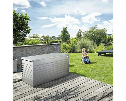 Graue Gartenbox auf einer Holzterrasse, im Hintergrund ein spielendes Kind und eine grüne Gartenlandschaft