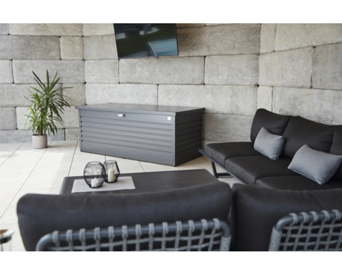 Außenbereich mit einer grauen Gerätebox, einer Couch, einem Tisch und einer Pflanze.