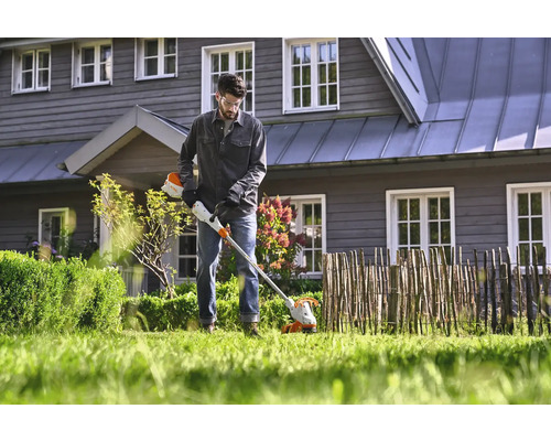 Mann arbeitet mit einem Akku-Rasentrimmer im Garten vor einem Haus.
