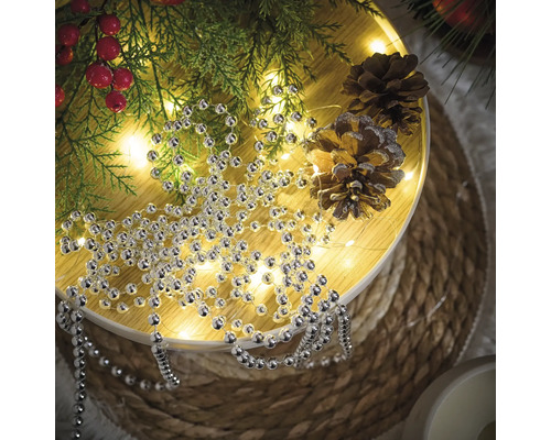 Dekoration mit Silberperlenkette, Tannenzweigen und Tannenzapfen auf einem runden Tisch.