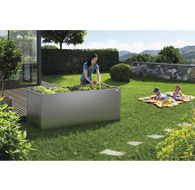 Szene im Garten mit einem erhöhten Beet aus Metall, einer Frau beim Ernten von Erdbeeren und spielenden Kindern im Hintergrund.