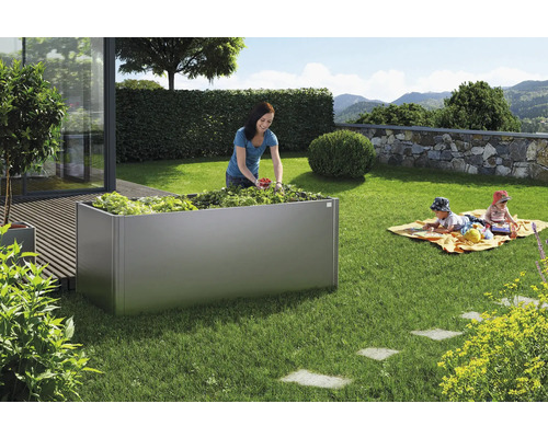 Szene im Garten mit einem Hochbeet aus Metall, einer Frau bei der Ernte und spielenden Kindern auf einer Decke.