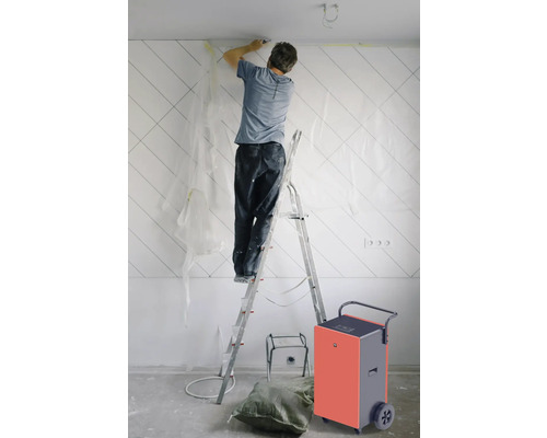 Ein Mann auf einer Leiter bei Renovierungsarbeiten mit einem Bautrockner im Raum.