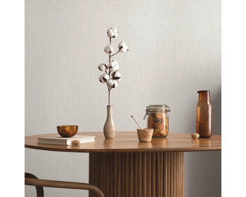 Tischdekoration mit Baumwollzweig, Schale, Buch, Vorratsglas und Flasche auf einem Holztisch vor einer einfarbigen Wand.