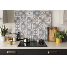 Organisierte helle Küche mit einer Rückwand mit grau-gelbem Fliesenmuster hinter einem Kochfeld auf weißer Arbeitsplatte mit Utensilien und Pflanzen.