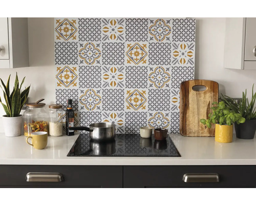 Organisierte helle Küche mit einer Rückwand mit grau-gelbem Fliesenmuster hinter einem Kochfeld auf weißer Arbeitsplatte mit Utensilien und Pflanzen.