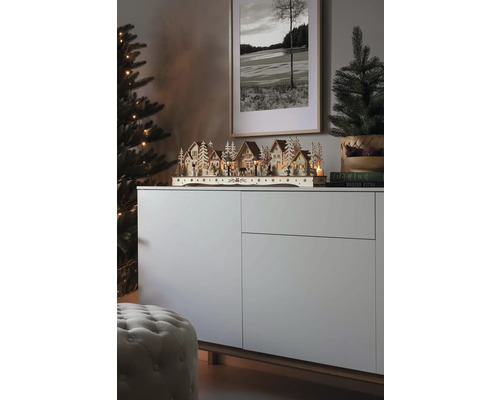 Weihnachtliche Szene mit einem beleuchteten Holzdorf auf einem Sideboard, neben einem Weihnachtsbaum und einem gerahmten Bild.