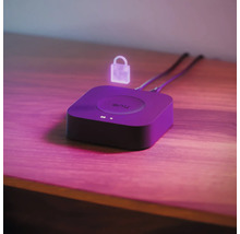Philips Hue Bridge auf einer Holzoberfläche mit einem schwebenden Symbol für Sicherheit und zwei angeschlossenen Kabeln.