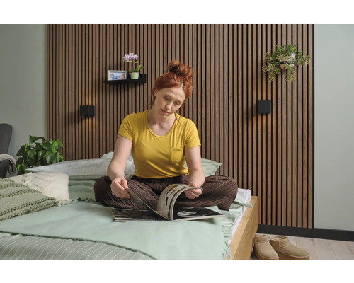 Eine Frau sitzt auf einem Bett vor einer Wandverkleidung und liest eine Zeitschrift. Das Schlafzimmer ist hell und freundlich eingerichtet.