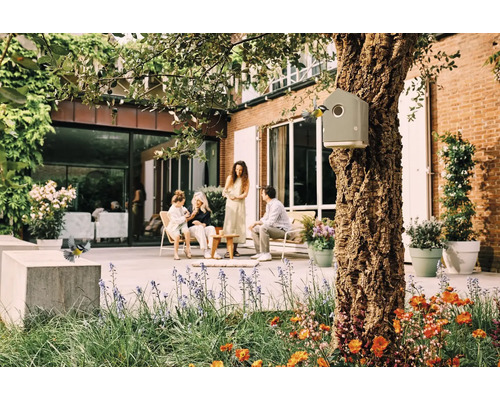 Szene im Garten mit Vogelhaus an einem Baum, sitzenden Personen und blühenden Blumen