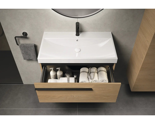 Waschtischunterschrank mit offenem Schubfach, darin Handtücher und Pflegeprodukte. Über dem Waschbecken hängt ein Spiegel.