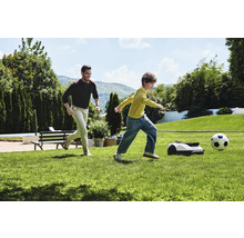 Mann und Kind spielen Fußball mit Rasenmähroboter im Garten.