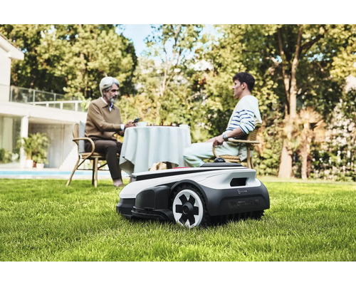 Szene mit einem Rasenmähroboter im Garten, während zwei Personen an einem Tisch sitzen