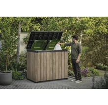 Mülltonnenbox aus Holz mit zwei grünen Mülltonnen und einer Person, die einen Müllsack hineinwirft