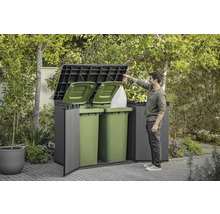 Mülltonnenbox im Garten mit zwei grünen Mülltonnen und einer Person, die einen Müllsack einwirft