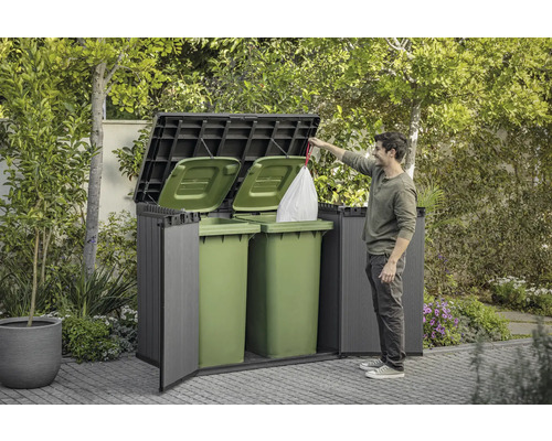 Mülltonnenbox im Garten mit zwei grünen Mülltonnen und einer Person, die einen Müllsack einwirft