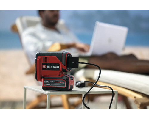 Einhell Akku-Lampe mit USB-Anschluss auf einem Tisch, im Hintergrund eine Person mit Laptop