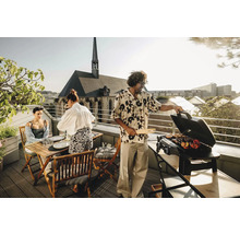 Personen grillen auf einer Dachterrasse mit Gasgrill und Holzzange, während im Hintergrund eine Kirche zu sehen ist.