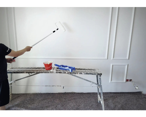 Maler streicht eine Wand mit einer Farbrolle, ein Farbeimer und eine Farbwanne stehen auf einem Gerüst.