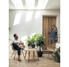 Heller Raum mit Mann im Sessel, Frau an Holztür, Tisch und Zimmerpflanzen.