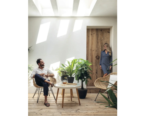 Heller Raum mit Mann im Sessel, Frau an Holztür, Tisch und Zimmerpflanzen.