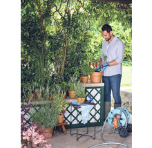 Ein Mann gießt im Garten Pflanzen in Töpfen, umgeben von Grün und einem Schlauchwagen.