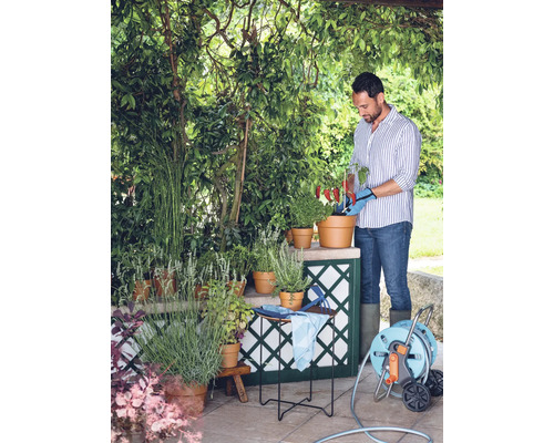 Ein Mann gießt im Garten Pflanzen in Töpfen, umgeben von Grün und einem Schlauchwagen.