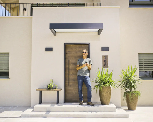 Moderner Eingangsbereich mit Haustür in Holzoptik, dunklem Vordach und Außenwandleuchten. Ein Mann steht vor der Tür und nutzt sein Smartphone.