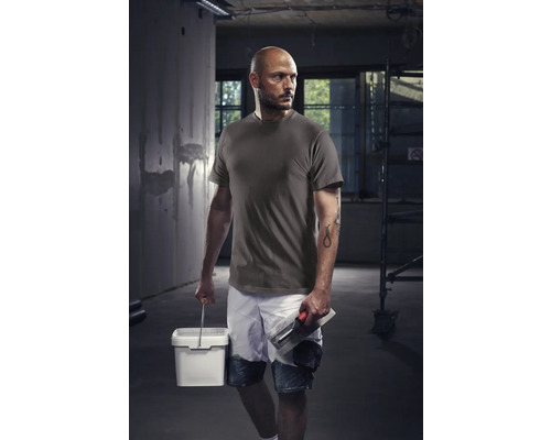 Handwerker mit T-Shirt, Shorts, Eimer und Glättkelle auf einer Baustelle.