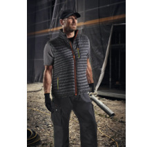 Mann trägt Arbeitskleidung mit Weste, T-Shirt, Hose und Mütze vor einer Baustelle