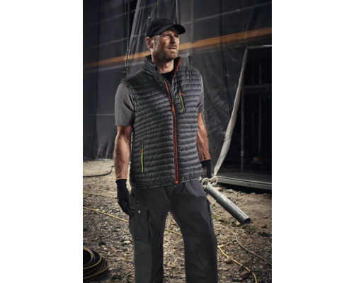 Mann trägt Arbeitskleidung mit Weste, T-Shirt, Hose und Mütze vor einer Baustelle